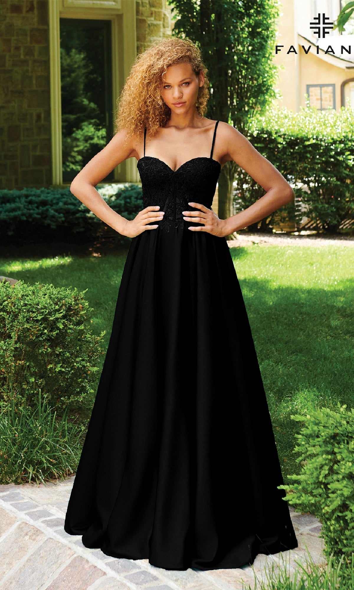 A woman stands outdoors on a stone path wearing the Faviana 11259 Lace-Up Long A-Line Prom Dress. Green shrubs and a building are visible in the background.